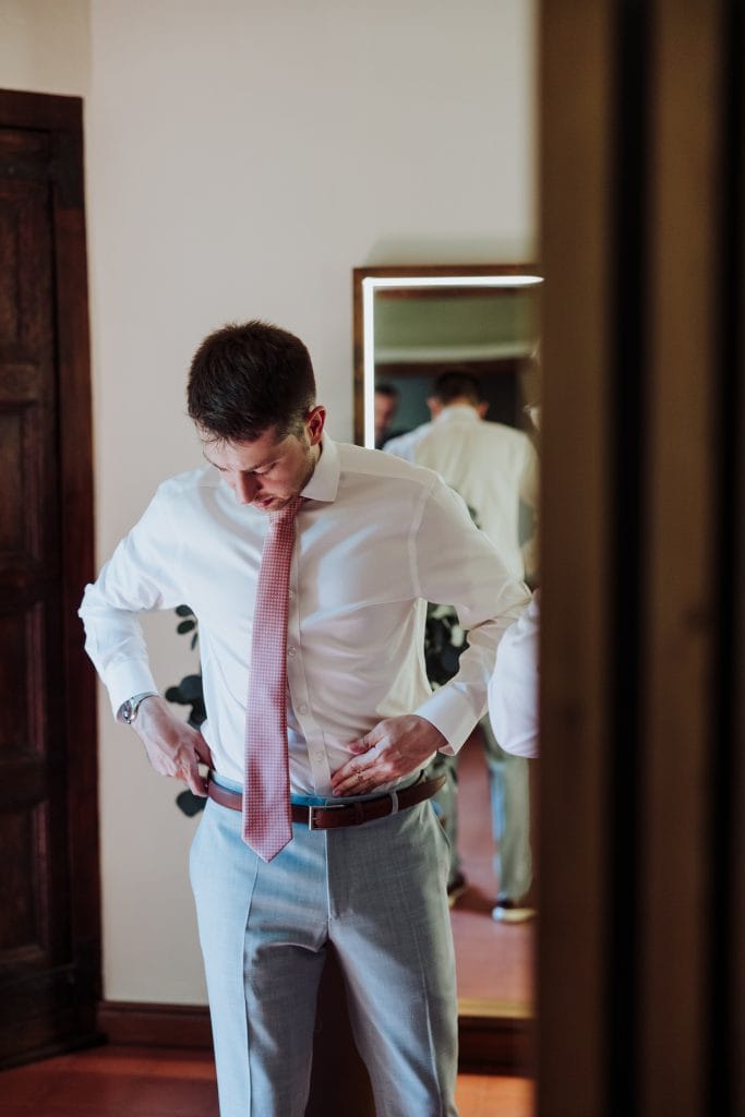Hombre ajustándose el cinturón en un espejo, vestido formal para una boda atípica.