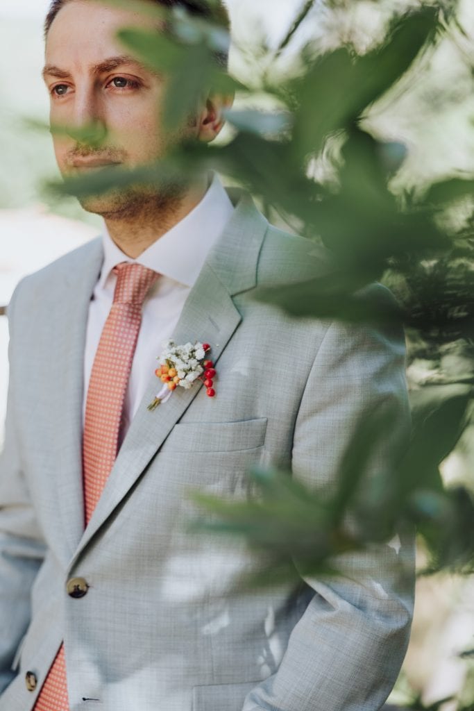 Novio en traje gris con flor en la solapa, rodeado de vegetación.