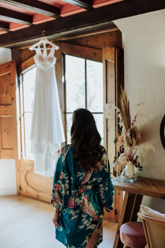 Vestido de novia colgado en habitación con ventanas de madera y decoración floral.