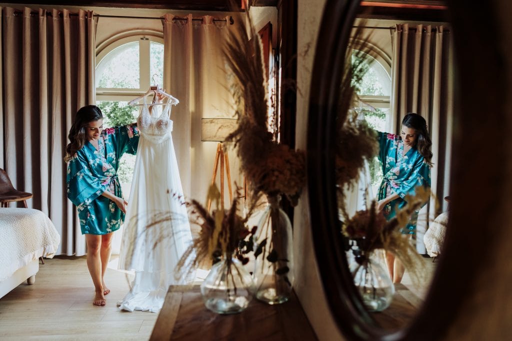 Vestido de novia en espejo, mujer en habitación con cortinas y decoración natural.