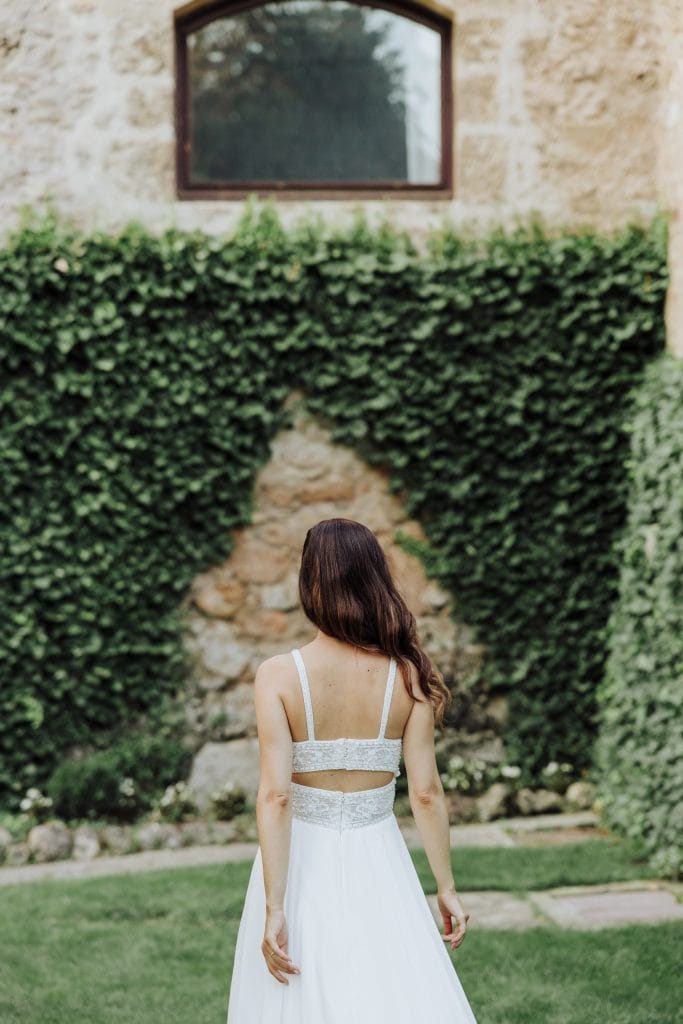 Vestido de novia con encaje y estilo bohemio en un jardín con paredes de piedra y vegetación.