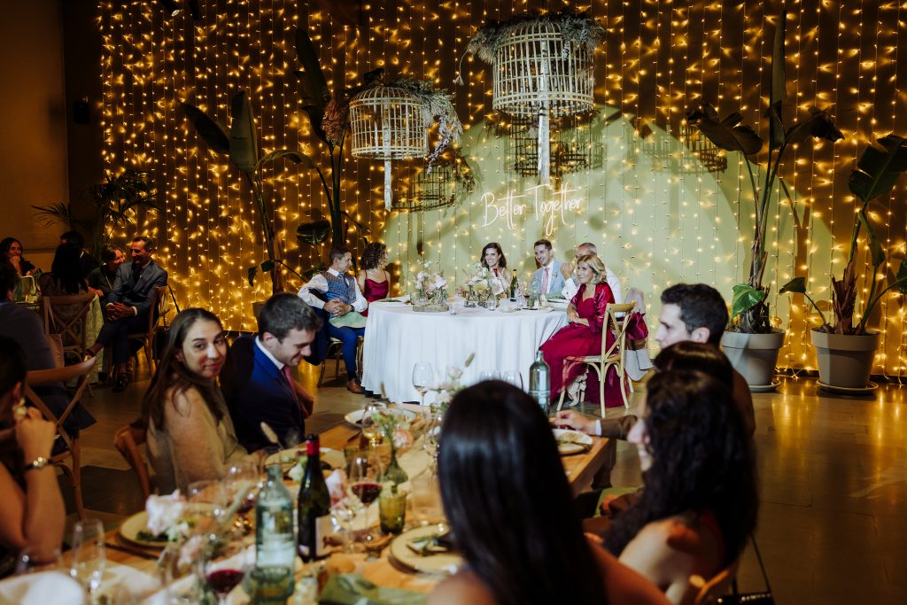 Boda en interior con decoración de luces cálidas y plantas, con invitados disfrutando de la ceremoni.
